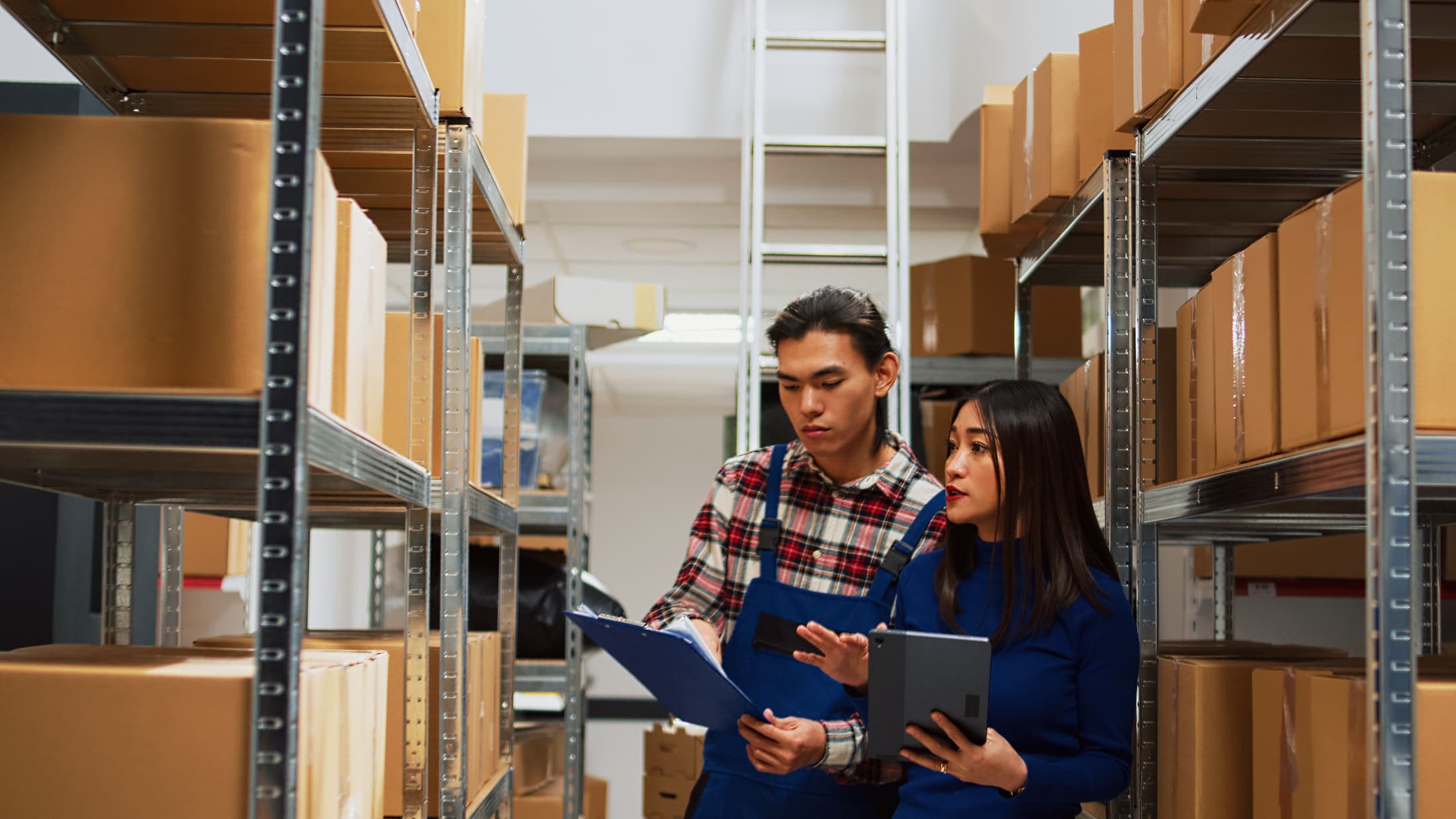 Two warehouse staff checking supplies against a tablet and clipboard down a racking aisle